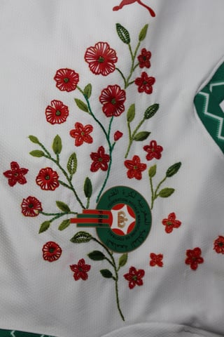 Embroidered floral design on white fabric featuring red flowers and green leaves with a decorative watering can