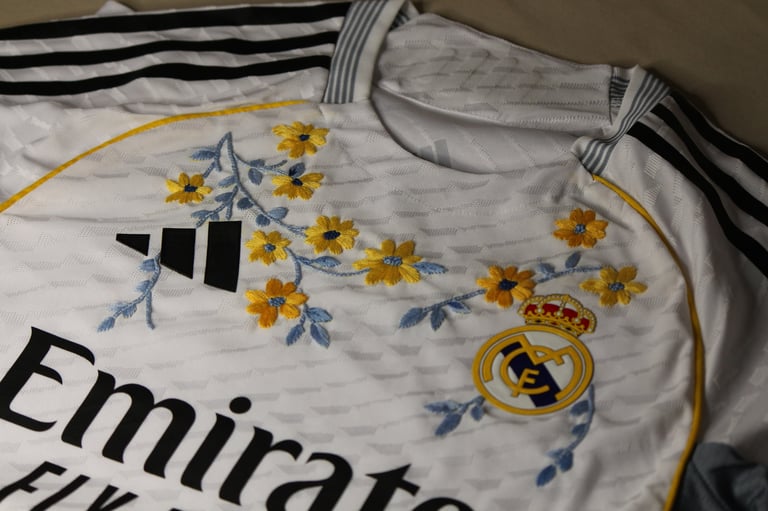 Real Madrid football jersey detail showing Adidas logo, Emirates sponsor text, and decorative floral embroidery with yellow flowers